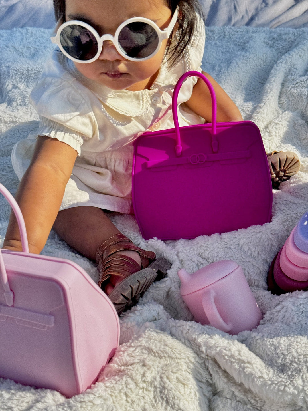 Child with sunglasses and pink and purple Birkkid Snack tote handbags on a sandy surface
