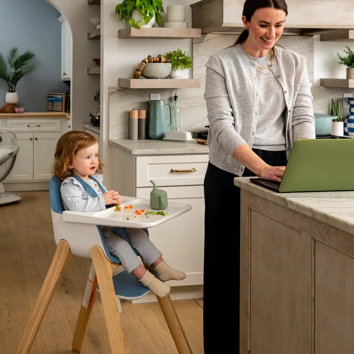 Woman using a laptop while a child sits in a n Uppababy Ciro high chair in a kitchen.