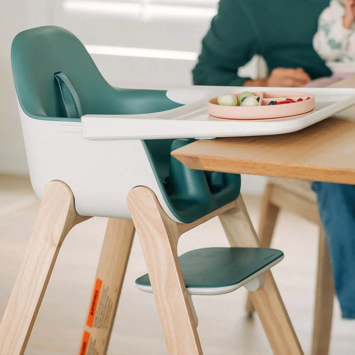 Children's high chair with wooden legs and green seat and backrest, placed at a dining table.