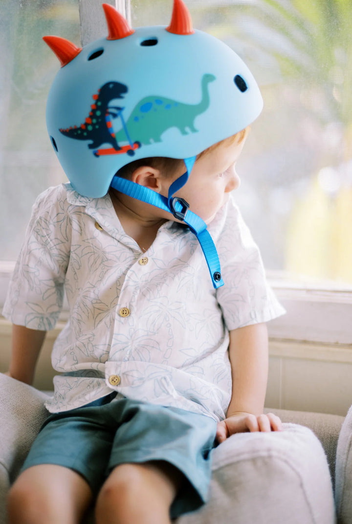 Child wearing a blue helmet with dinosaur design sitting on a couch.