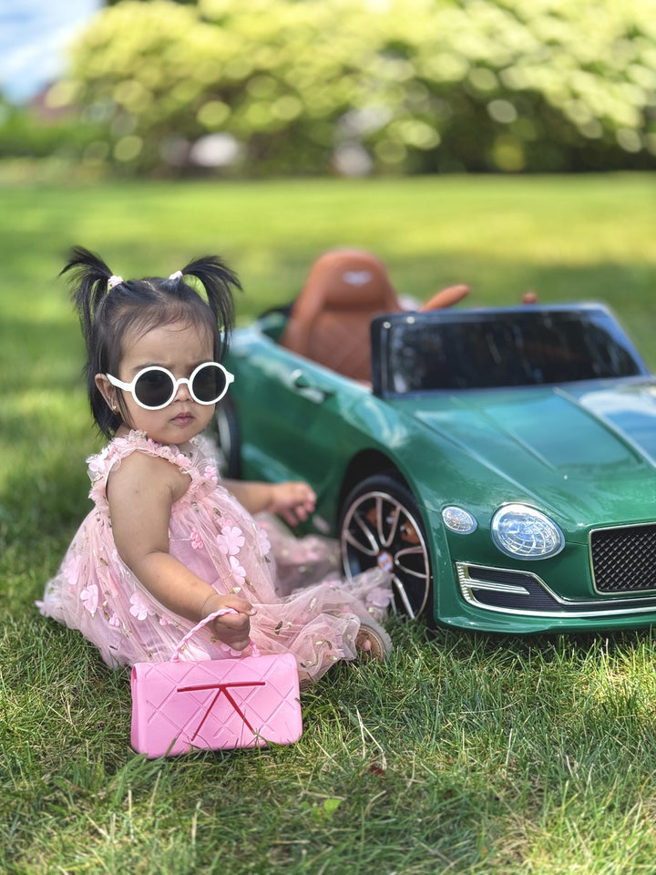 Child in a pink dress and sunglasses sitting next to a green toy car on grass whole holding a Chanellini snack tote handbag by Moonfare