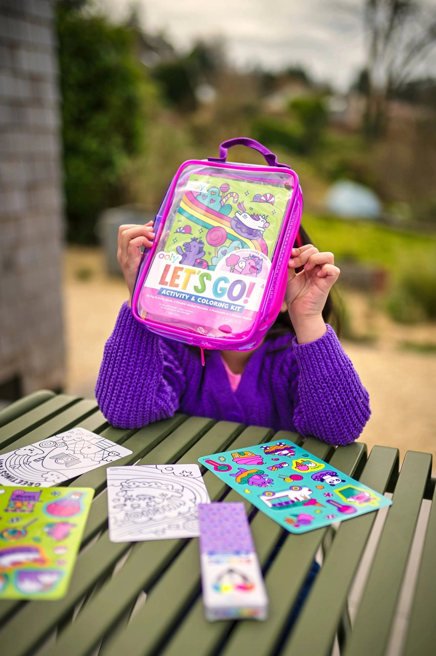 Child holding a colorful activity kit with 'Let's Go!' text, surrounded by coloring books and stickers on a green table outdoors.