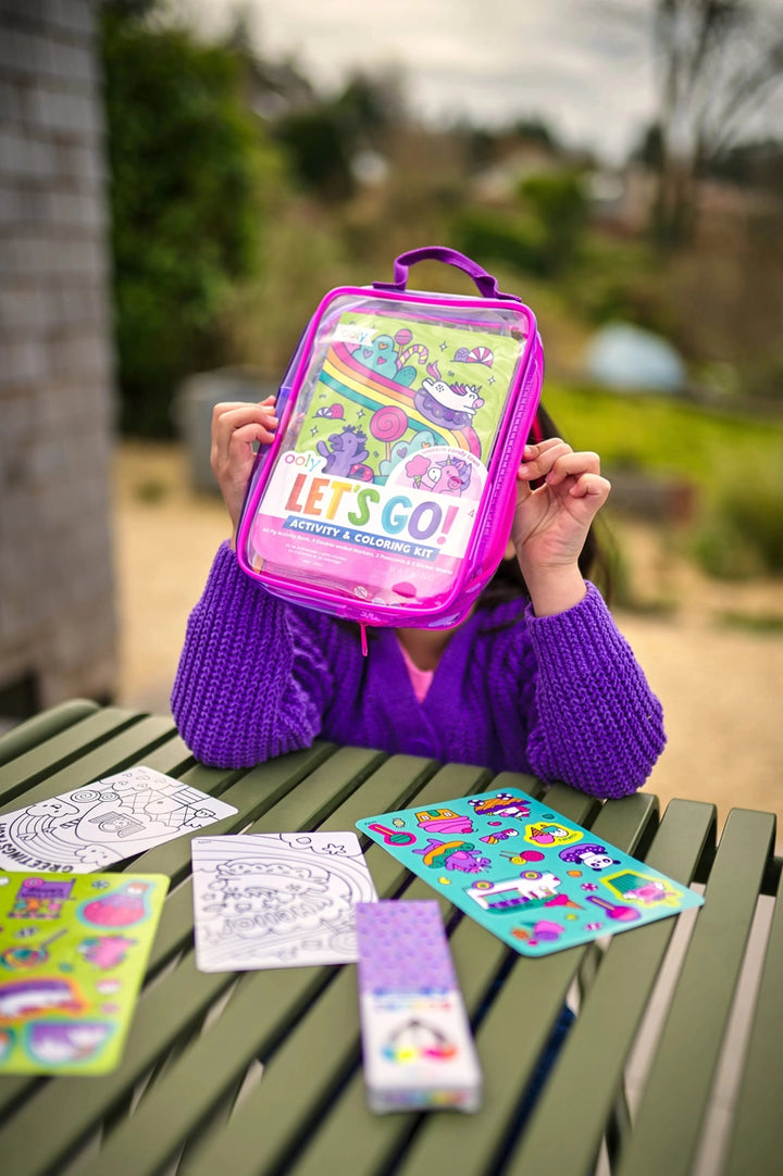 Child holding a colorful activity kit with 'Let's Go!' text, surrounded by coloring books and stickers on a green table outdoors.