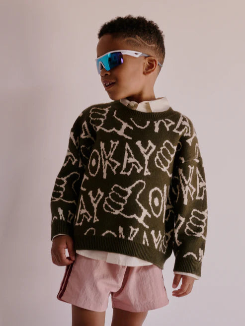 Children’s oversized crewneck sweater in pink with hand-drawn ‘okay’ lettering and thumbs-up doodle, laid flat to show boxy fit and ribbed trim