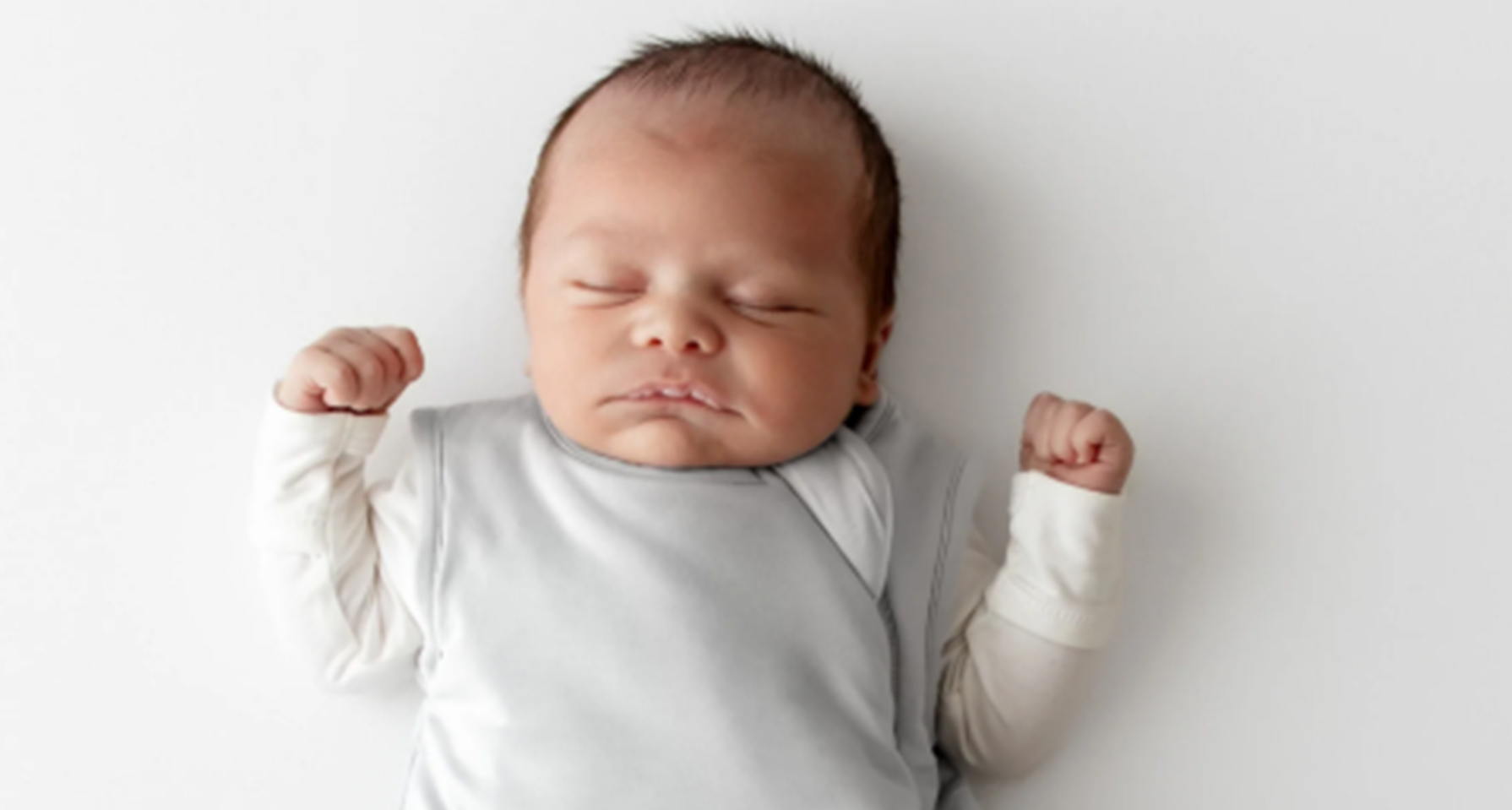 A sleeping newborn baby lying on their back, swaddled in a light gray blanket with long sleeves and white cuffs, against a plain white background.