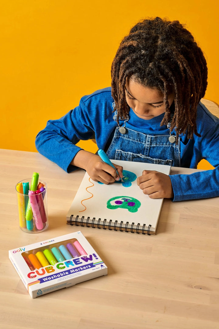 Child drawing with markers on a notebook at a table, with a box of Crayola markers in the foreground.