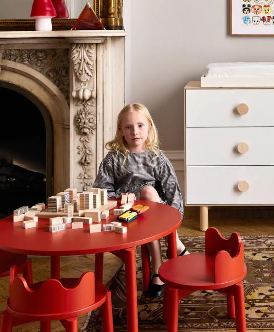 Child playing with wooden blocks at a red table in a room with a white dresser and decorative fireplace.