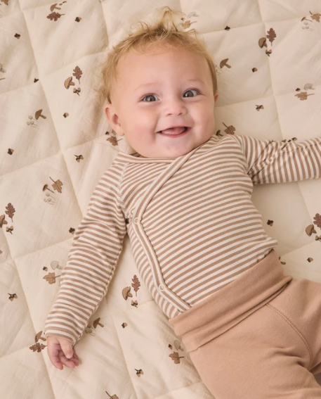 A smiling baby lying on a quilted cream mattress with a brown leaf pattern, wearing a beige and white striped long-sleeve wrap bodysuit and tan pants.