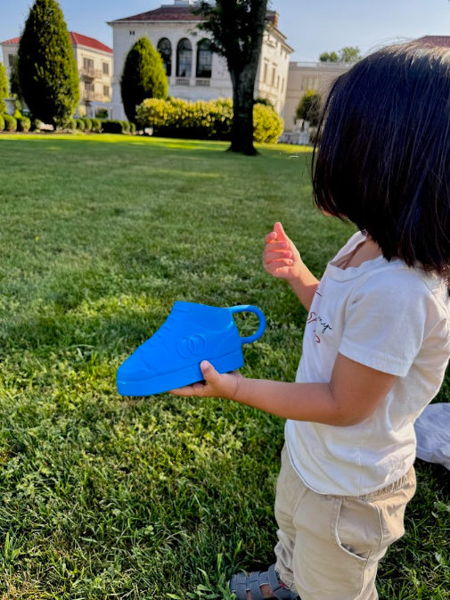 Child holding a blue Moon force sneaker snack tote in a grassy area with a building in the background