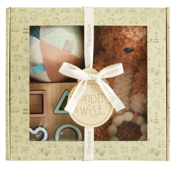 Wonder & Wise baby gift set in a cream-colored patterned box with a clear window, tied with a white ribbon. Visible contents include a brown teddy bear, a wooden shape toy, and soft baby accessories.