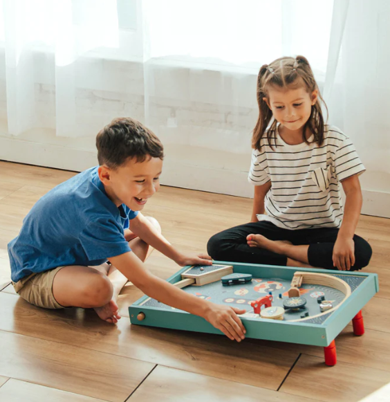 Two children playing with a wooden toy train set on a light wood floor in a bright room. The toy set is placed on a mint green tray with small red legs.