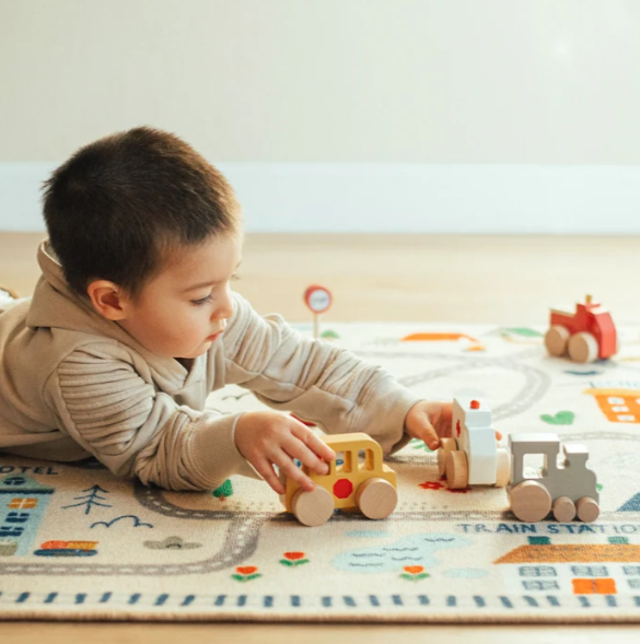 A young child lies on a colorful play rug, playing with wooden toy trains in a bright indoor room.